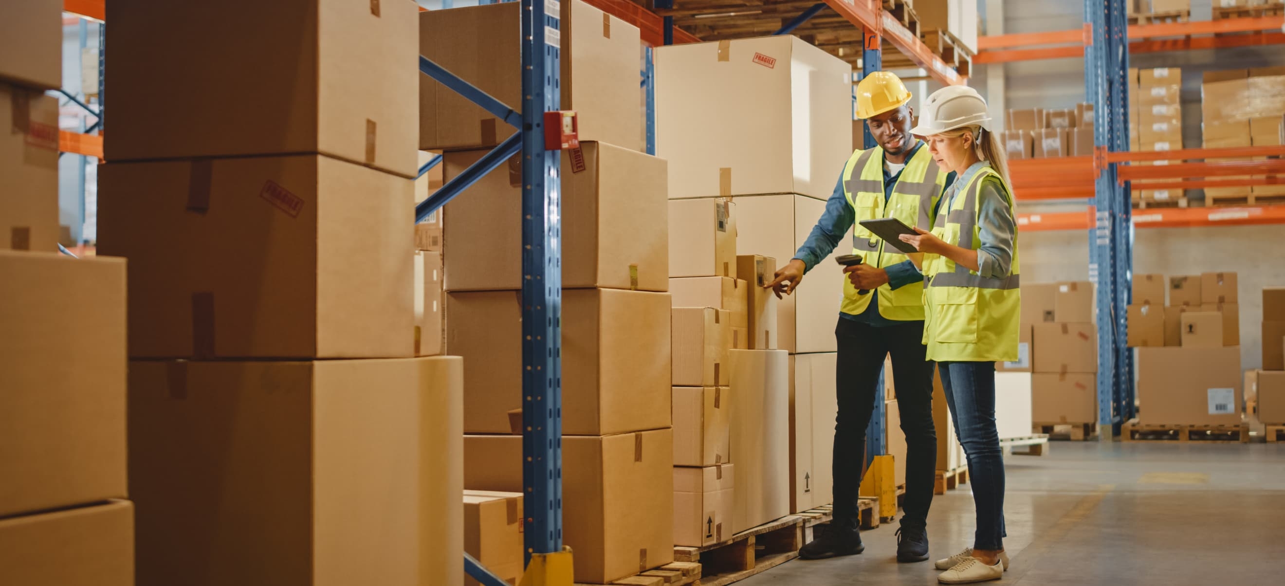 Two workers wearing safety vests and helmets are inspecting stacked boxes in a warehouse. One holds a tablet. They are surrounded by shelves filled with more boxes.