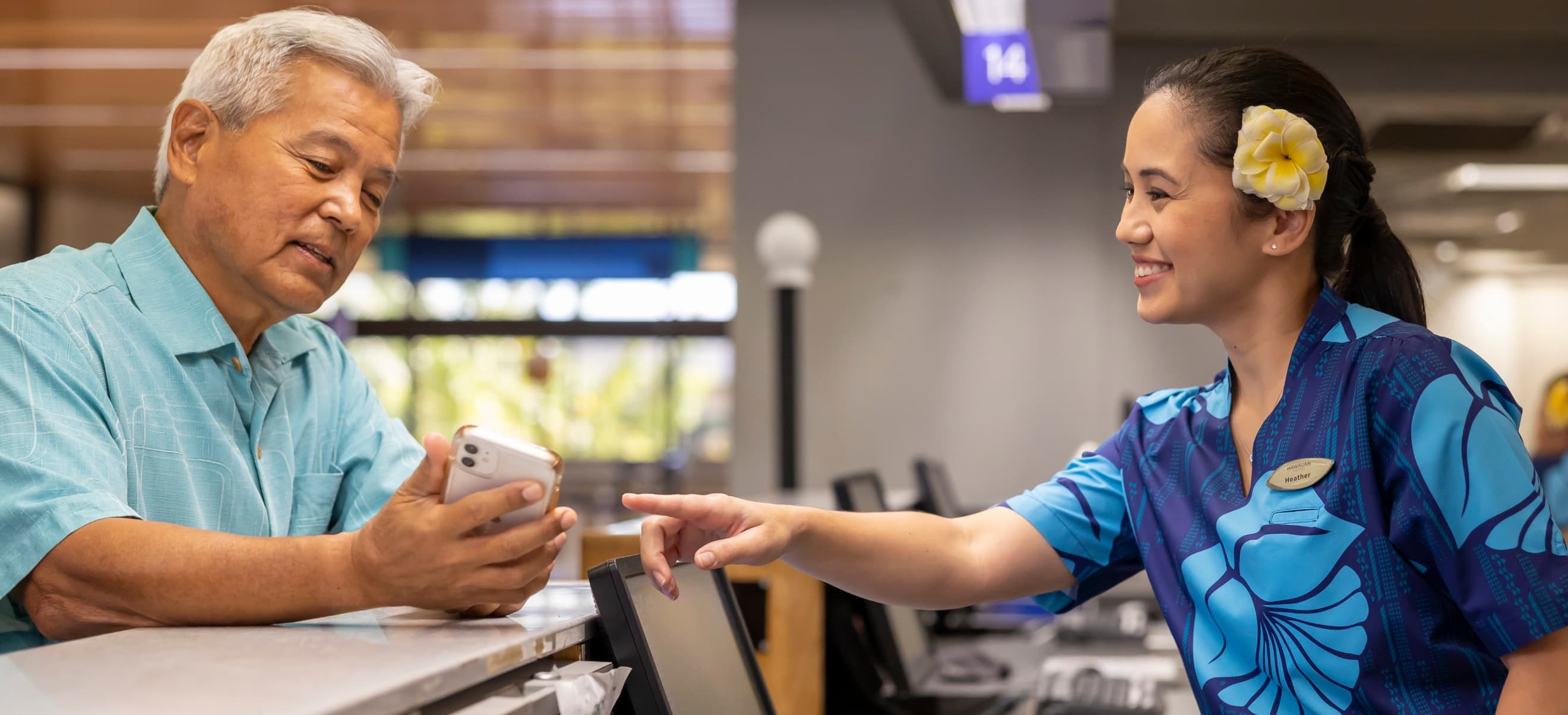 An elderly man in a blue shirt shows his Hawaiian Airlines app by ArcTouch to a smiling woman at a counter. The woman, wearing a blue uniform and flower in her hair, points to the phone.