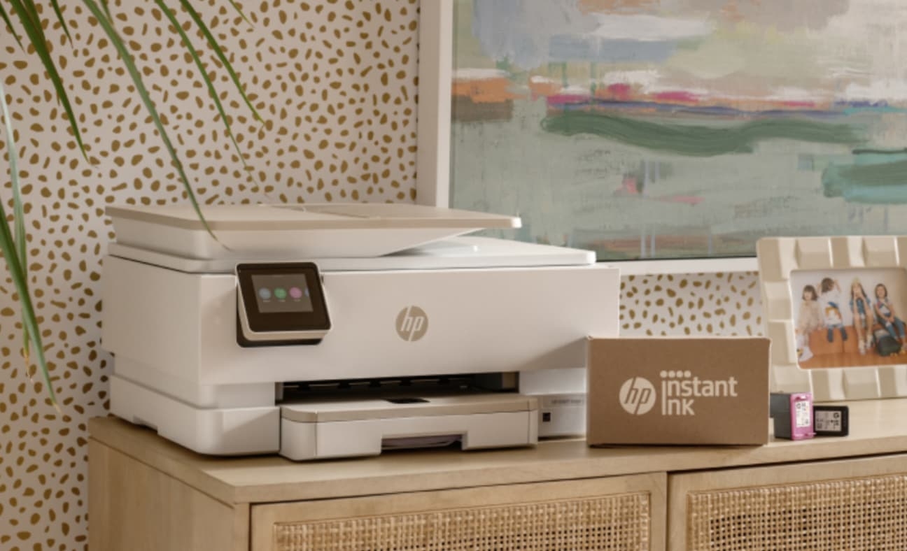 A white HP printer is on a wooden cabinet beside a cardboard box labeled "HP Instant Ink app" and ink cartridges, reflecting the tools of a successful company case study by ArcTouch.