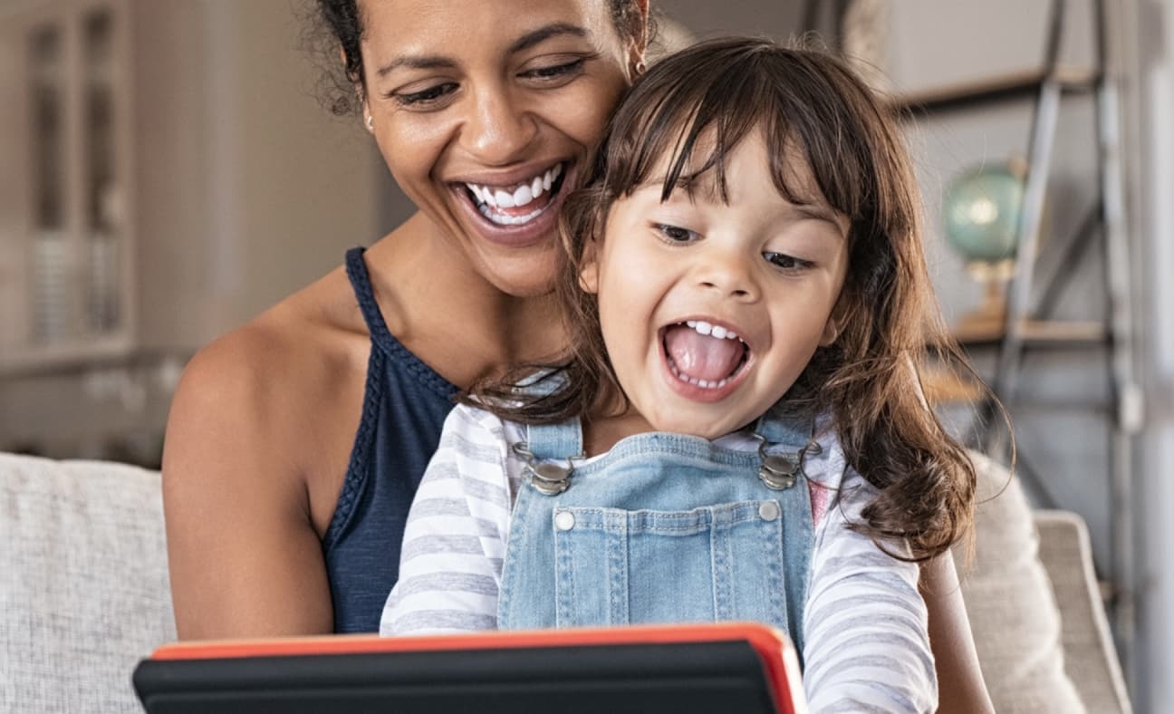 A woman and child sitting on a couch, smiling and looking at a tablet. Bright, colorful graphics adorn the image. They are enjoying ArcTouch's Common Sense Media app.
