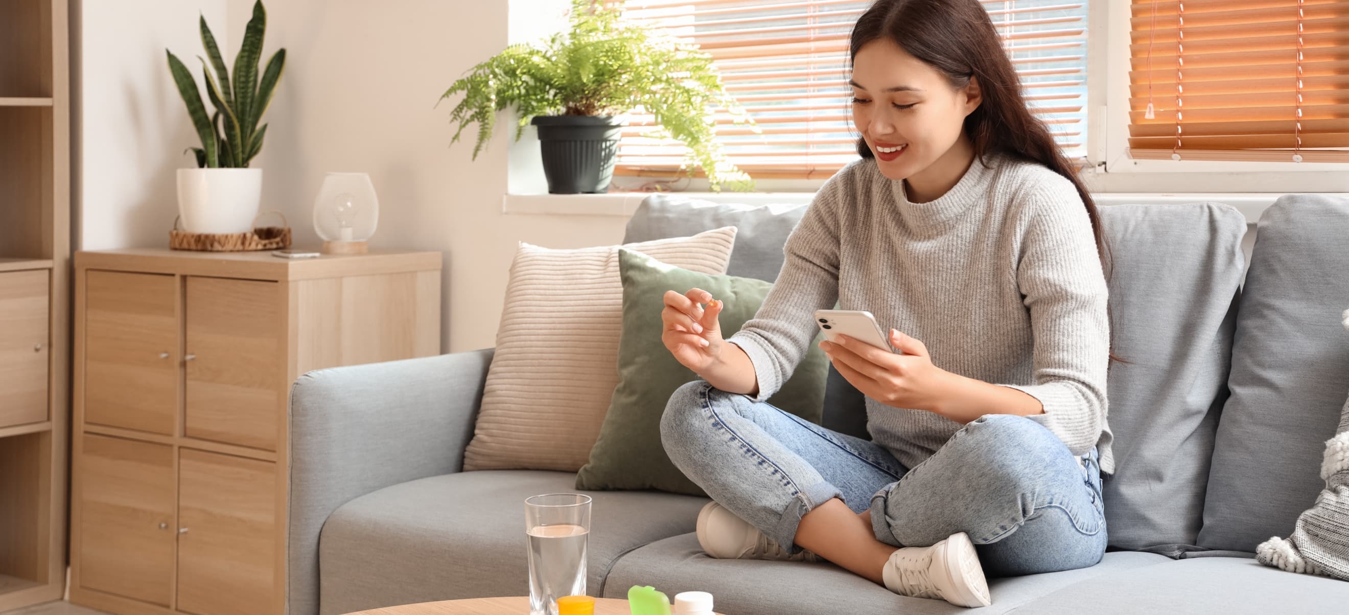 A woman sits cross-legged on a grey sofa, holding a smartphone with the Magellan app open and smiling. She is wearing a grey sweater and jeans. A glass of water and some items are on the table in front of her.