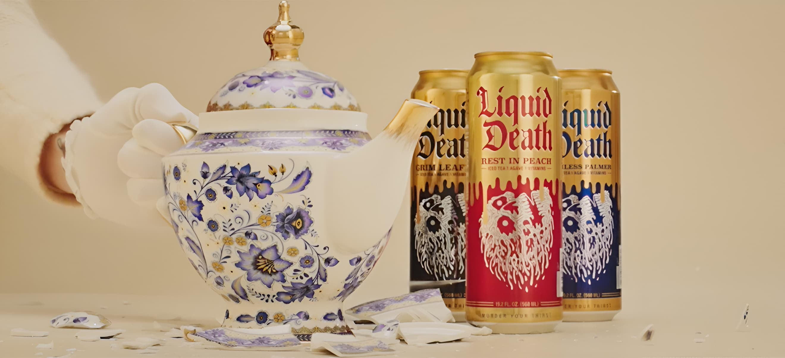 A hand wearing a white glove is holding a floral-patterned teapot. The teapot is broken, with pieces scattered below. Behind the teapot, there are two cans of Liquid Death.