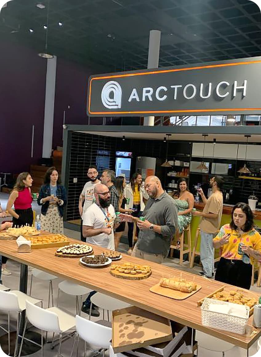 A group of people are gathered in a modern office setting with a table filled with various pastries and snacks. A sign with the text "ARCTOUCH" is prominently displayed above them.