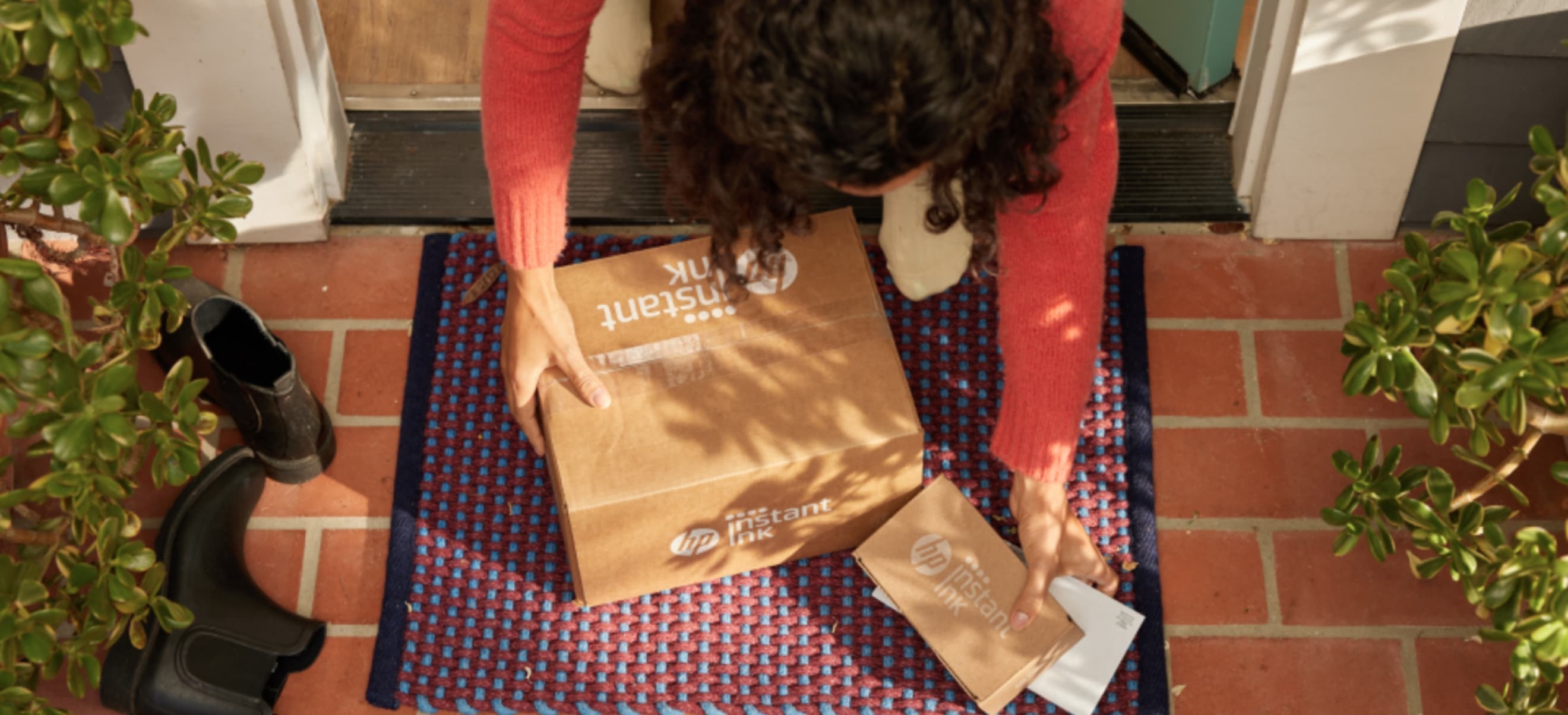 A person is kneeling on a doorstep, picking up two packages from the HP Instant Ink, next to a pair of black rain boots.