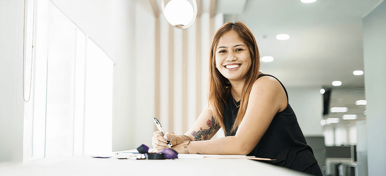 A woman with long hair, wearing a black sleeveless top, is smiling and sitting at a desk, writing with a pen. The background shows a well-lit modern interior.