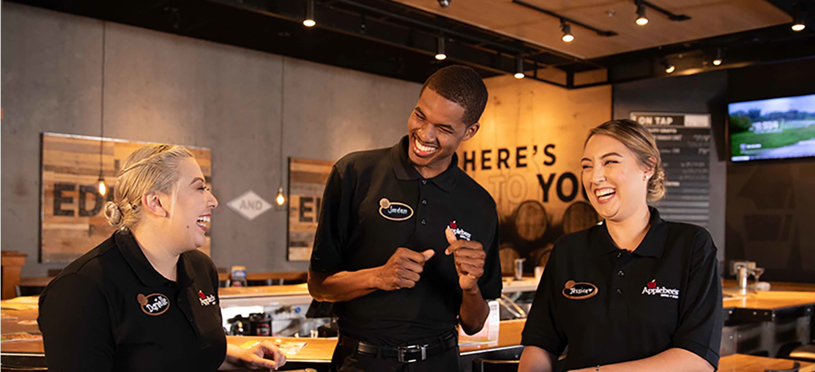 Three Applebee's employees in uniform, each wearing a name tag, stand behind a counter, smiling and engaging in conversation inside a modern restaurant.