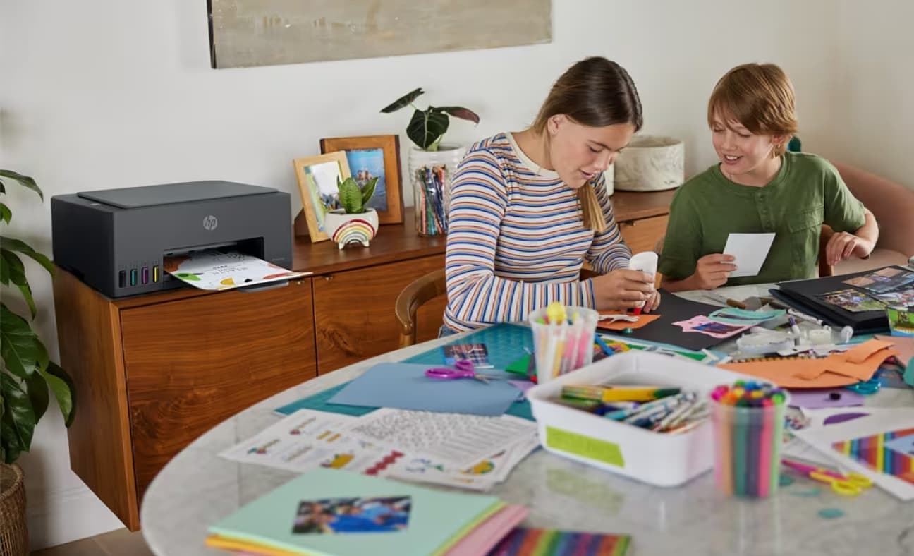 Two children are crafting at a table covered with supplies, while a printer on a wooden desk behind them prints photos using the HP Printables app.