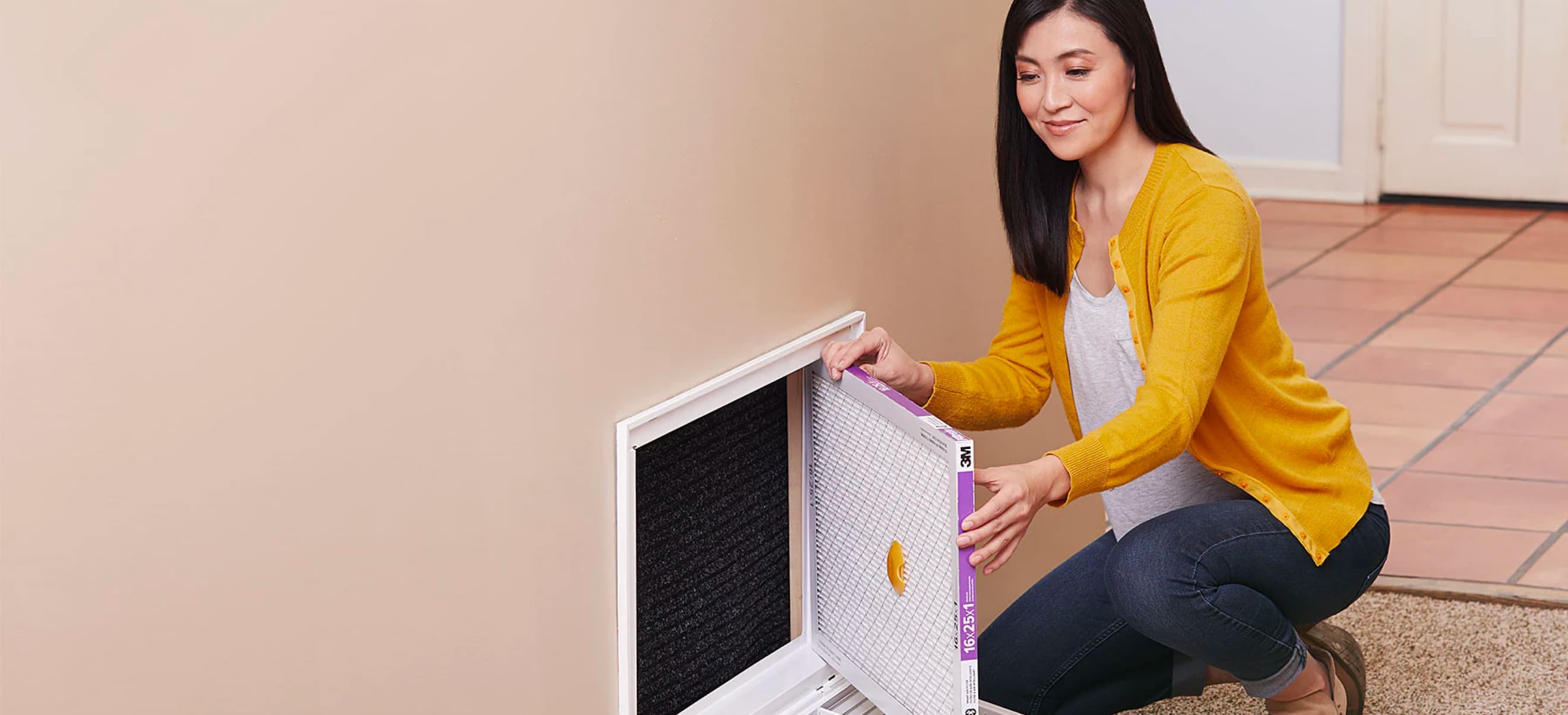 A woman is kneeling on the floor, installing a 3M Filtrete into a wall-mounted air return vent inside a home. She is wearing a yellow cardigan and blue jeans.
