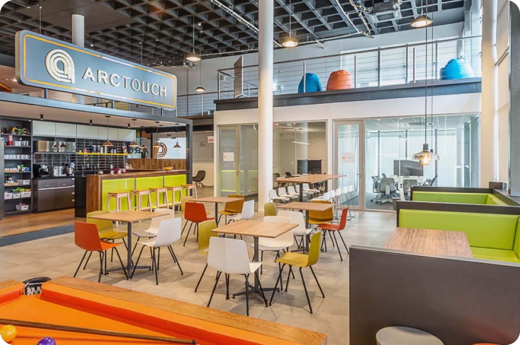 ArcTouch's office features a modern space with a kitchen area, various colored chairs and tables, and a pool table in the foreground. A large sign overhead reads "ARCTOUCH." An upper level with bean bags is also visible.