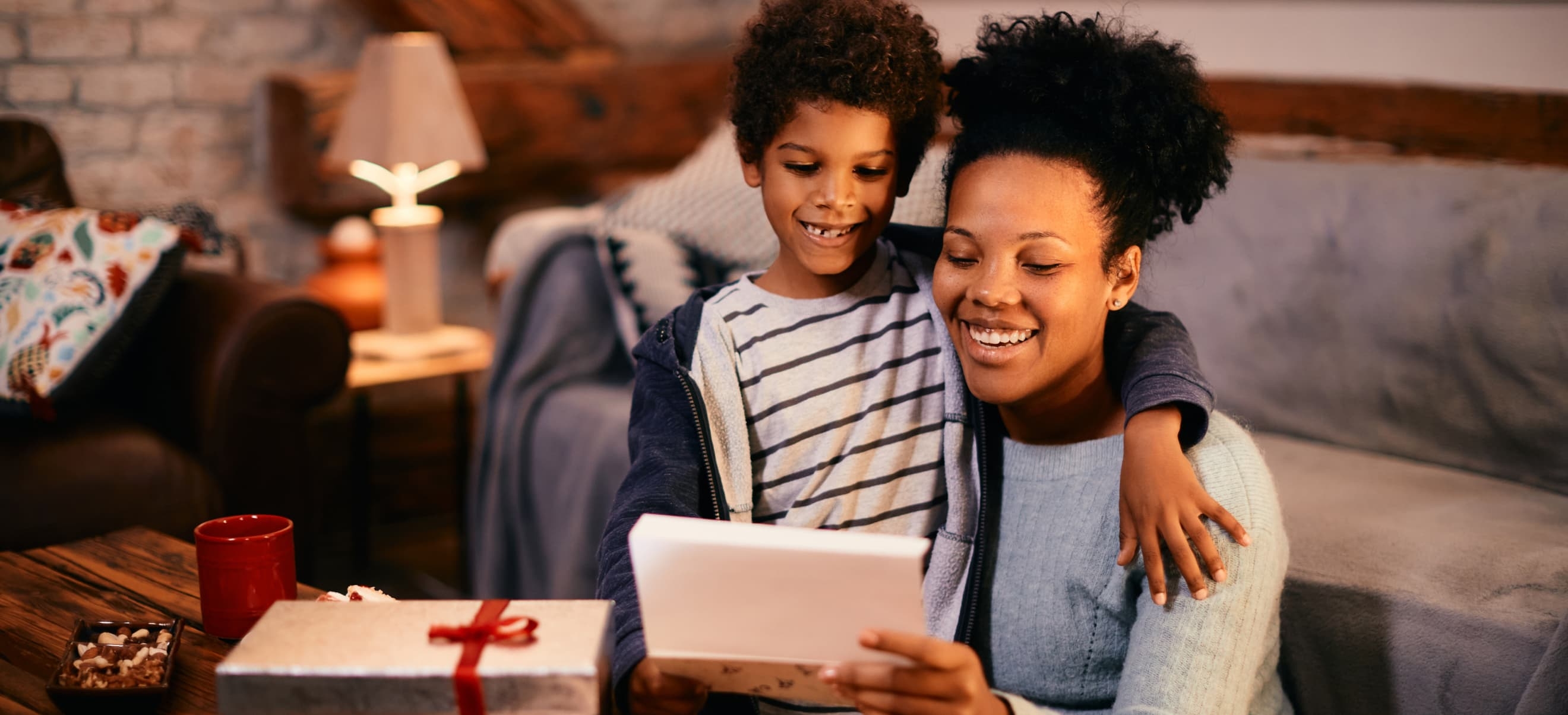 A woman and a child sit together on a couch, smiling while looking at a card. A wrapped gift and a red mug are on the table in front of them. A lit lamp is in the background, with the HP Printables app open on their tablet.