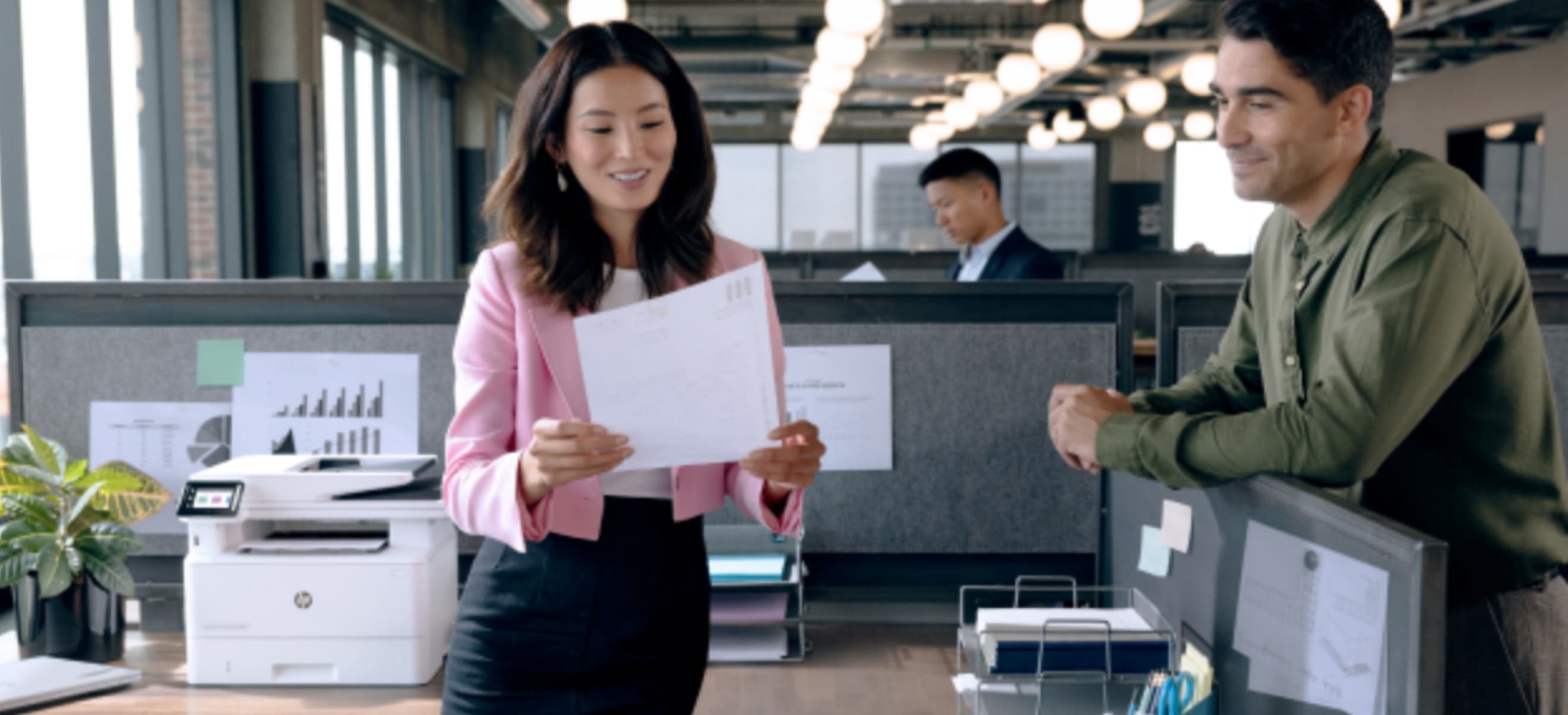 A woman holding an HP Instant Ink prints stands next to a man leaning on a cubicle in an office space with another person working at a desk in the background.