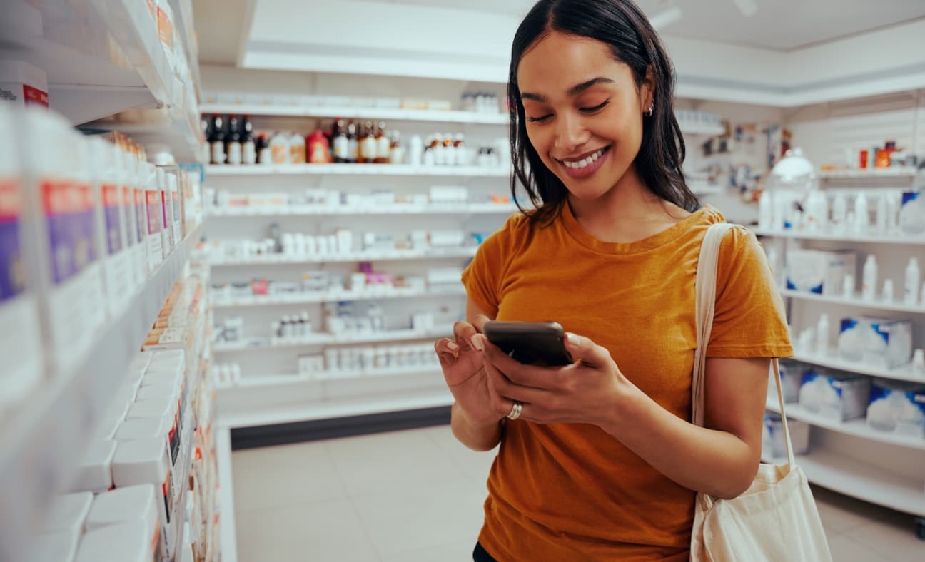 A woman in a pharmacy smiles as she looks at her smartphone. Shelves filled with various medicinal products are visible in the background, and she is reading a Magellan app case study.
