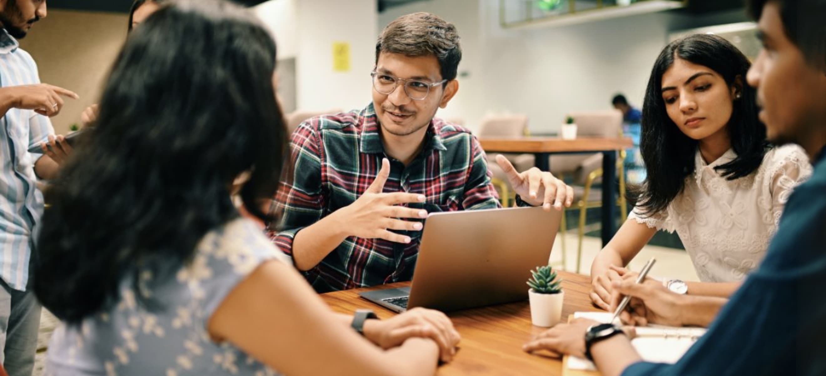 A group of four young adults are gathered around a table, engaged in discussion. One person is speaking and gesturing with hands, while others listen and take notes. A laptop and a small plant are on the table.