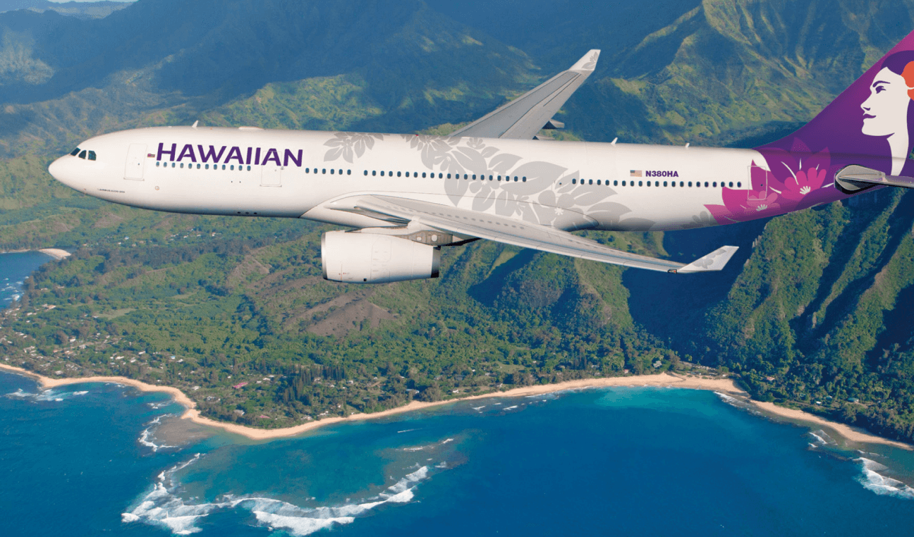 A Hawaiian Airlines airplane flies over a scenic coastline with lush green mountains and blue ocean waters
