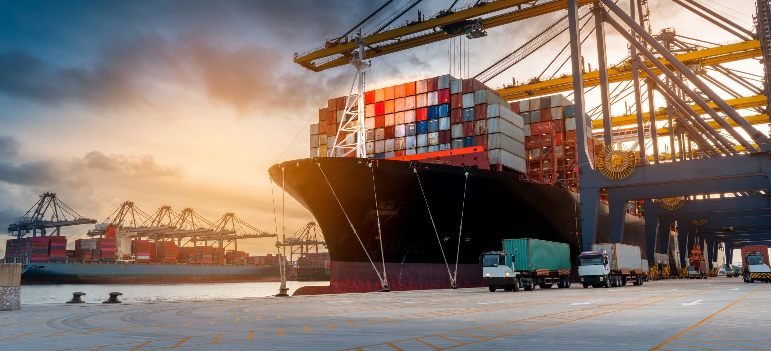 A large cargo ship docked at a port, with numerous colorful containers stacked on it. Cranes and trucks are visible in the background under a partly cloudy sky.