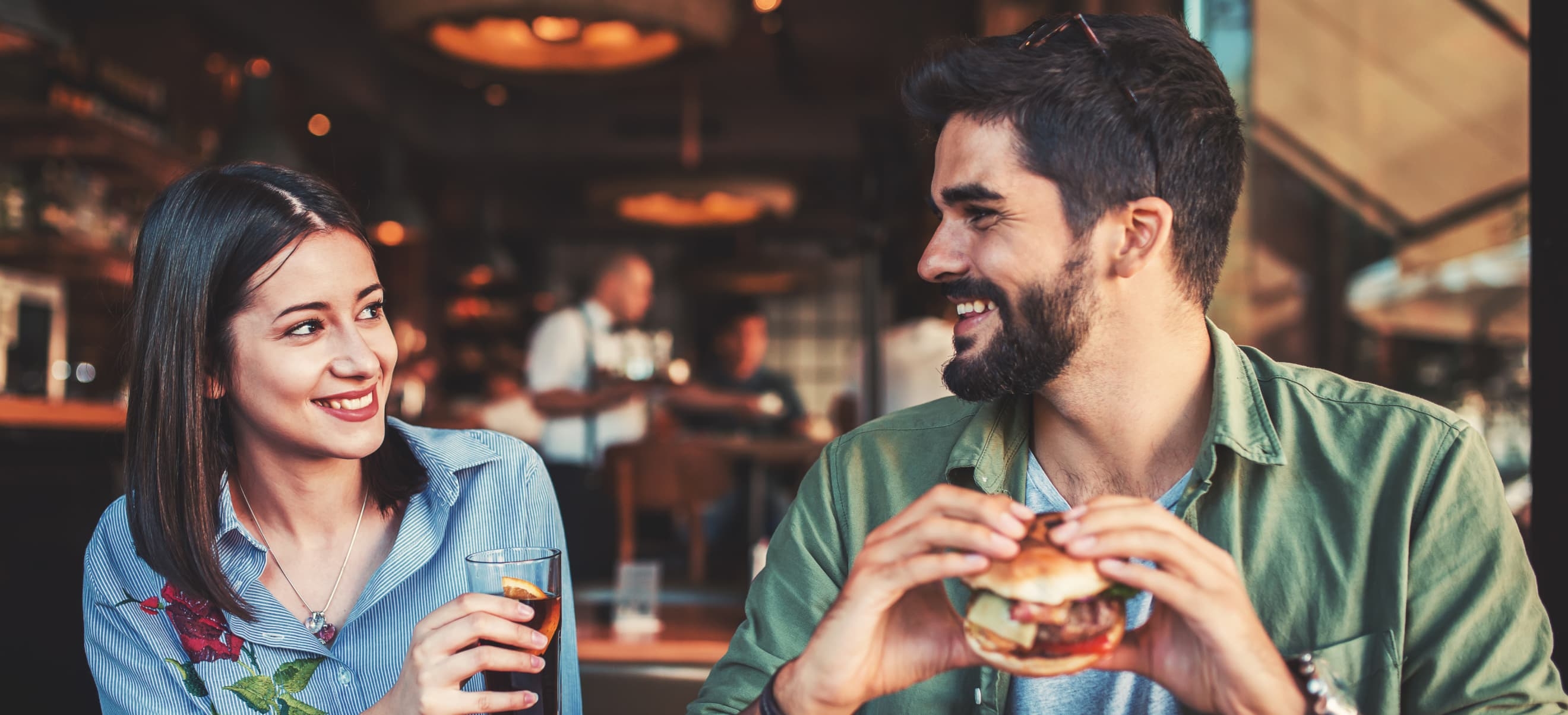 A woman and man are sitting at an Applebee's restaurant. The woman is holding a drink, smiling at the man, who is holding a burger. The background shows a bar with another person in the distance.