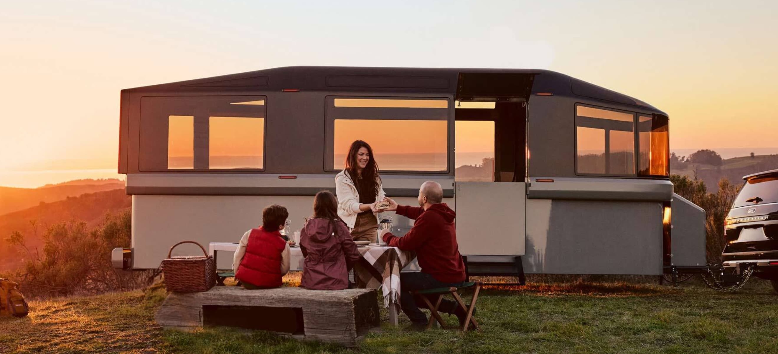 Four people are sitting around a picnic table, enjoying a meal in front of a large camper at sunset while using the Lightship RV app by ArcTouch.