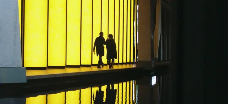 Photo displaying the silhouettes of two people walking across a luminous yellow structure on the front of a building.