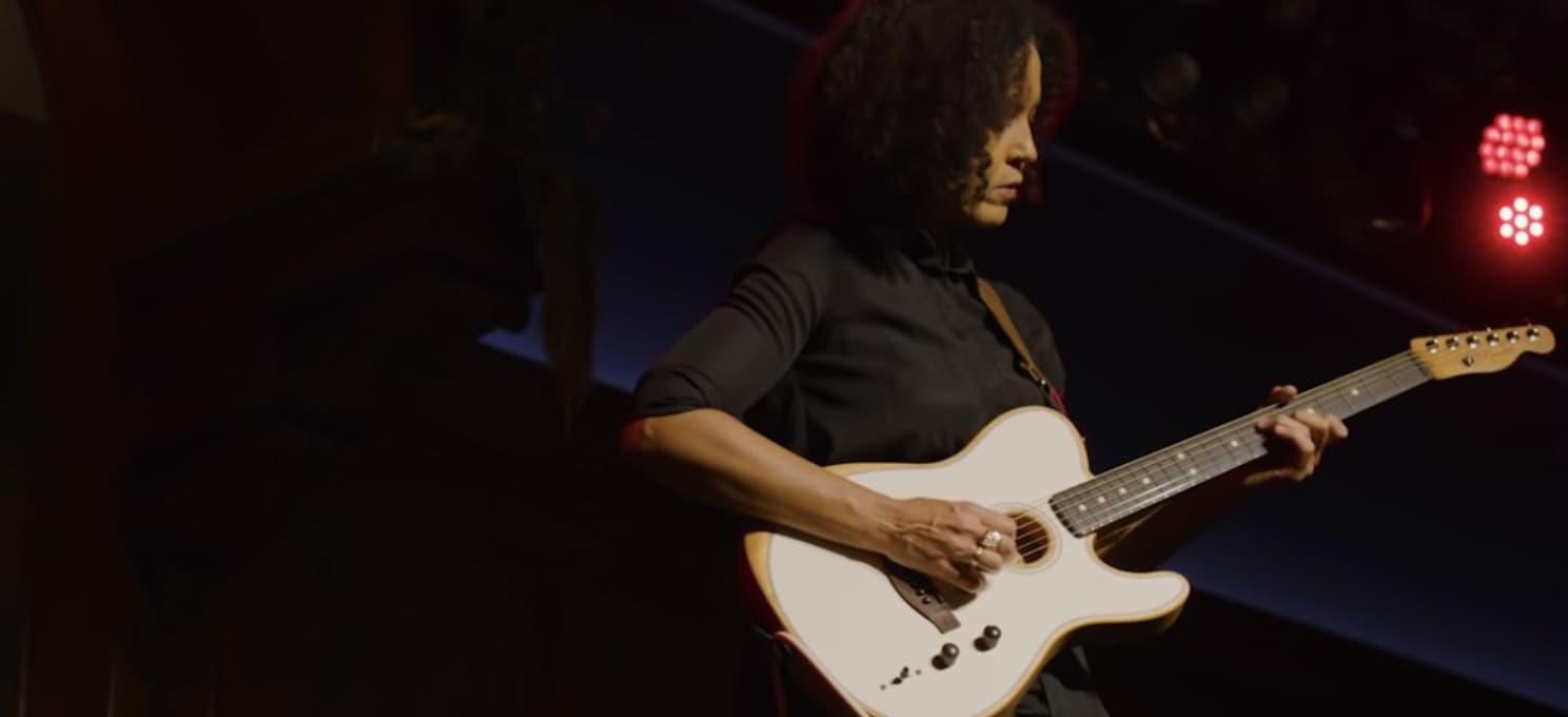 A person is playing a Fender guitar on stage under red lighting.