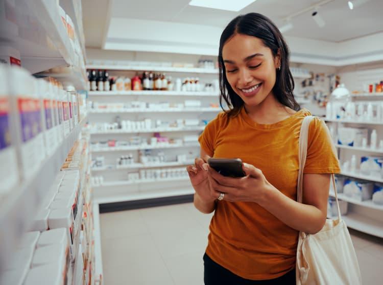 A woman in a pharmacy smiles as she looks at her smartphone. Shelves filled with various medicinal products are visible in the background, and she is reading a Magellan app case study.