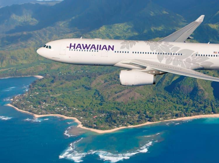 A Hawaiian Airlines airplane flies over a scenic coastline with lush green mountains and blue ocean waters