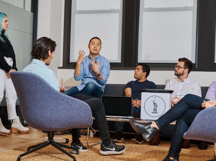 A group of people sits and stands in an office setting, engaged in a discussion. One person is speaking animatedly, while others listen attentively. An IBM Careers' website created by ArcTouch is open on a laptop placed on a table.