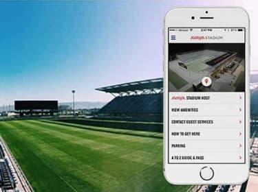 View of Avaya Stadium's interior with empty stands and a green field. An iPhone in the foreground displays the Avaya Stadium mobile app, efficiently built using Xamarin by ArcTouch, showcasing options for navigating the stadium.