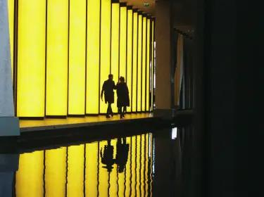 Photo displaying the silhouettes of two people walking across a luminous yellow structure on the front of a building.