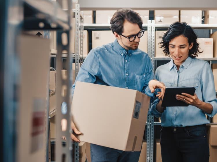 Two people in a storage area, both wearing denim shirts. One is holding a cardboard box while the other points at a tablet, possibly discussing inventory or logistics on the Cure website by ArcTouch.