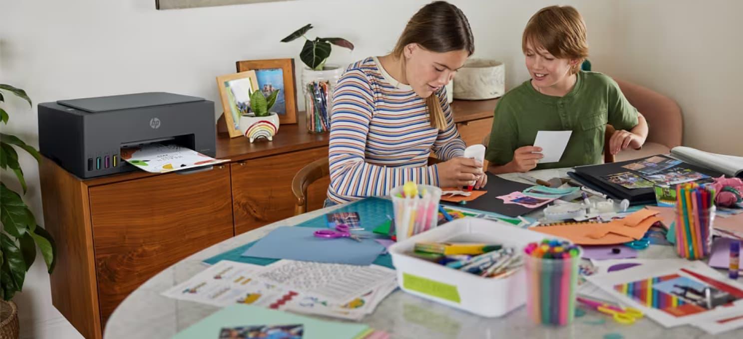 Two children are crafting at a table covered with supplies, while a printer on a wooden desk behind them prints photos using the HP Printables app.