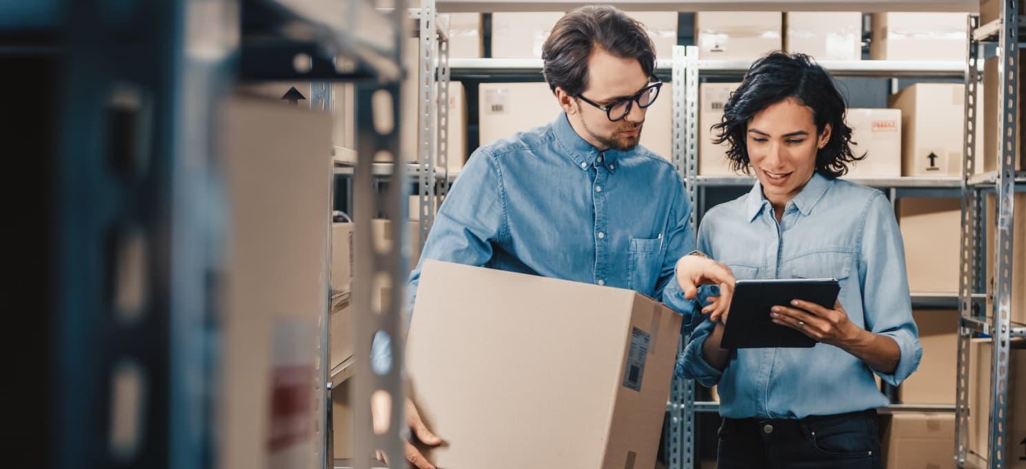 Two people in a storage area, both wearing denim shirts. One is holding a cardboard box while the other points at a tablet, possibly discussing inventory or logistics on the Cure website by ArcTouch.