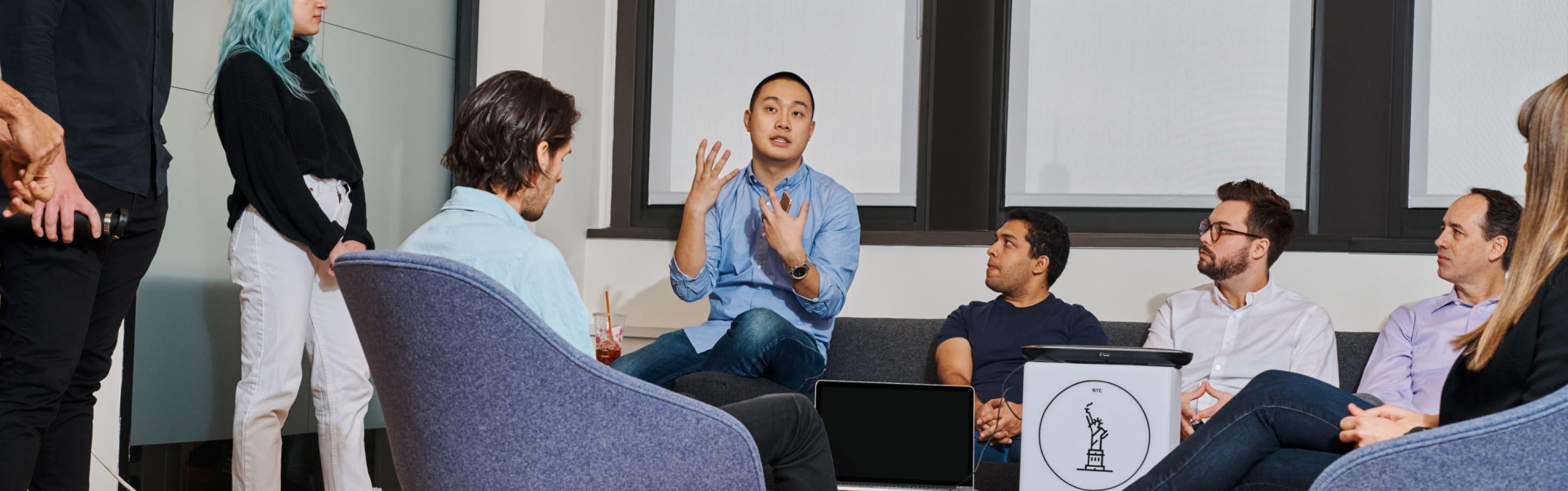 A group of people sits and stands in an office setting, engaged in a discussion. One person is speaking animatedly, while others listen attentively. An IBM Careers' website created by ArcTouch is open on a laptop placed on a table.