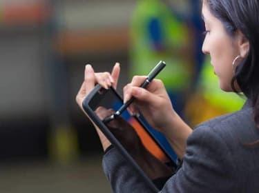 A woman in a gray blazer uses a stylus to write on a tablet, building excellence in mobile apps with every stroke against the blurred background.