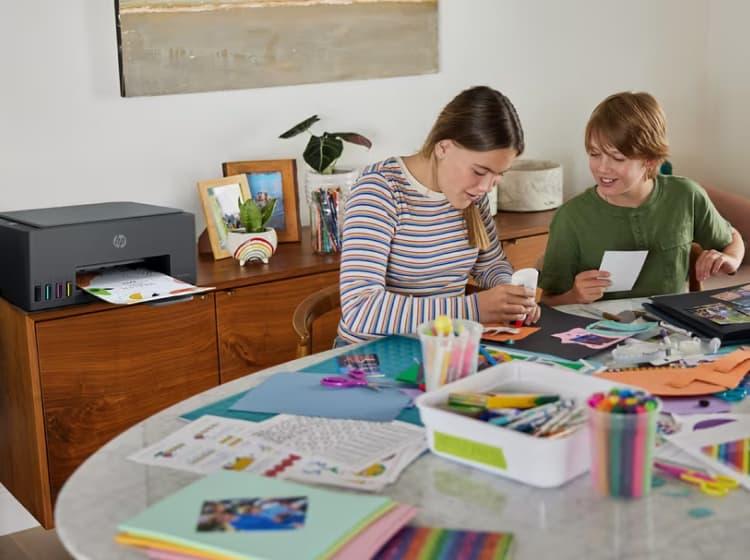 Two children are crafting at a table covered with supplies, while a printer on a wooden desk behind them prints photos using the HP Printables app.