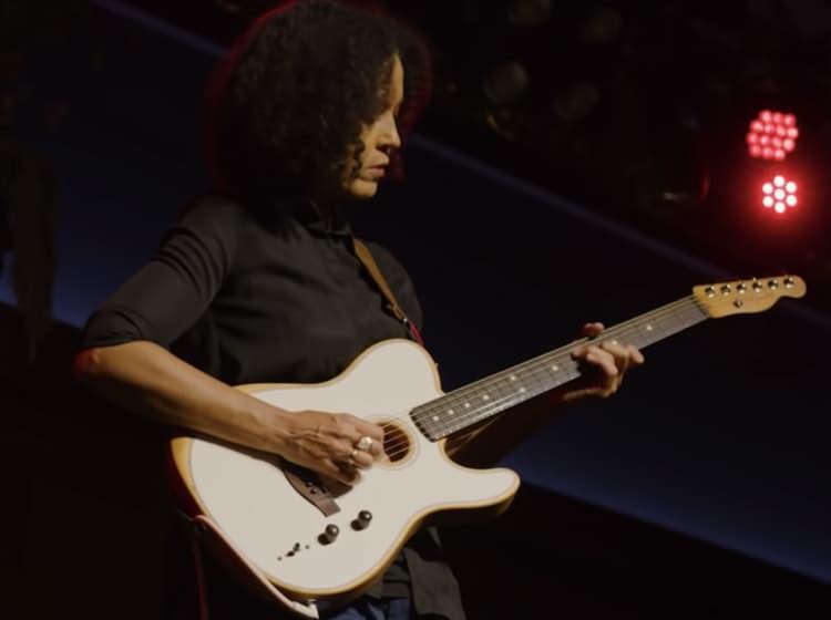 A person is playing a Fender guitar on stage under red lighting.