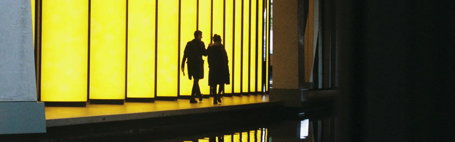 Photo displaying the silhouettes of two people walking across a luminous yellow structure on the front of a building.