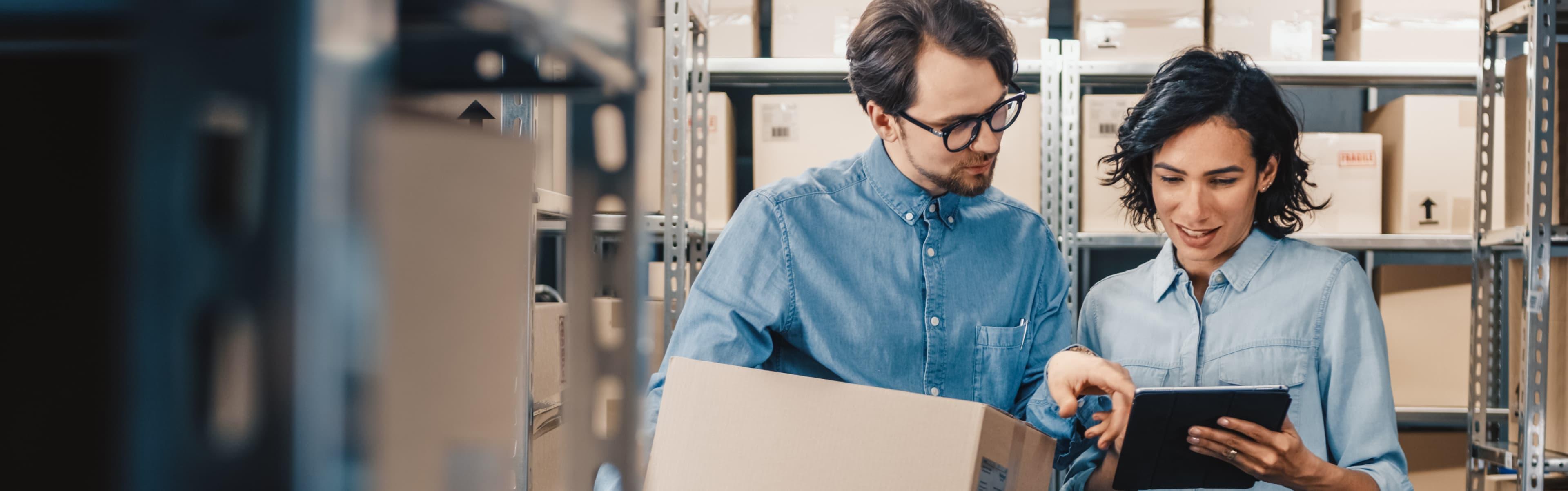 Two people in a storage area, both wearing denim shirts. One is holding a cardboard box while the other points at a tablet, possibly discussing inventory or logistics on the Cure website by ArcTouch.