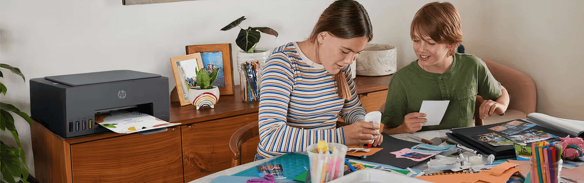 Two children are crafting at a table covered with supplies, while a printer on a wooden desk behind them prints photos using the HP Printables app.