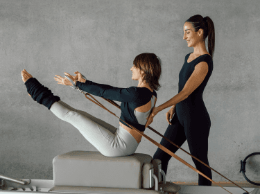 A woman performs a seated exercise using the Pilates Metrics healthcare app while an instructor stands beside her, providing guidance.