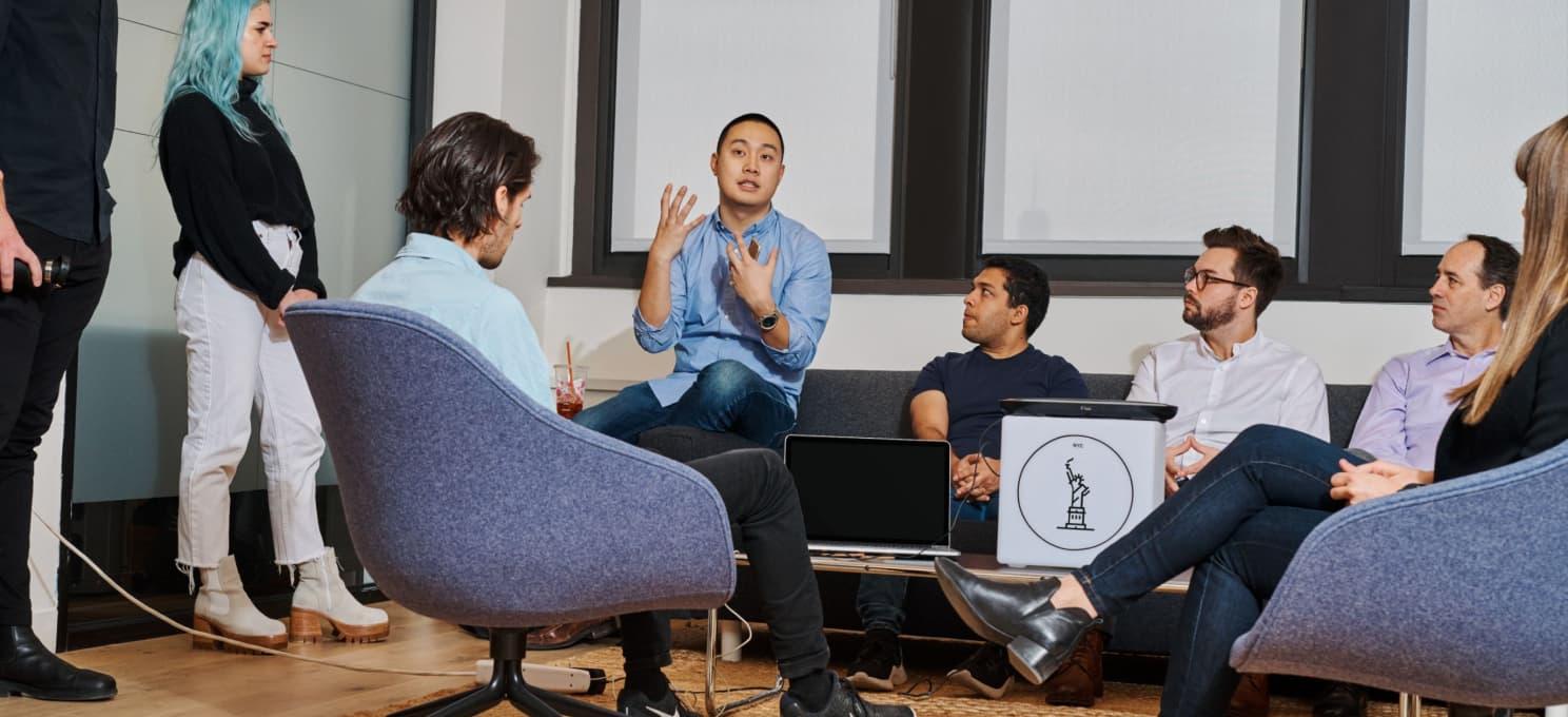 A group of people sits and stands in an office setting, engaged in a discussion. One person is speaking animatedly, while others listen attentively. An IBM Careers' website created by ArcTouch is open on a laptop placed on a table.