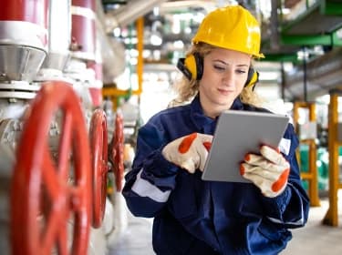 A woman dressed in protective work clothing operates a tablet in an industrial environment.