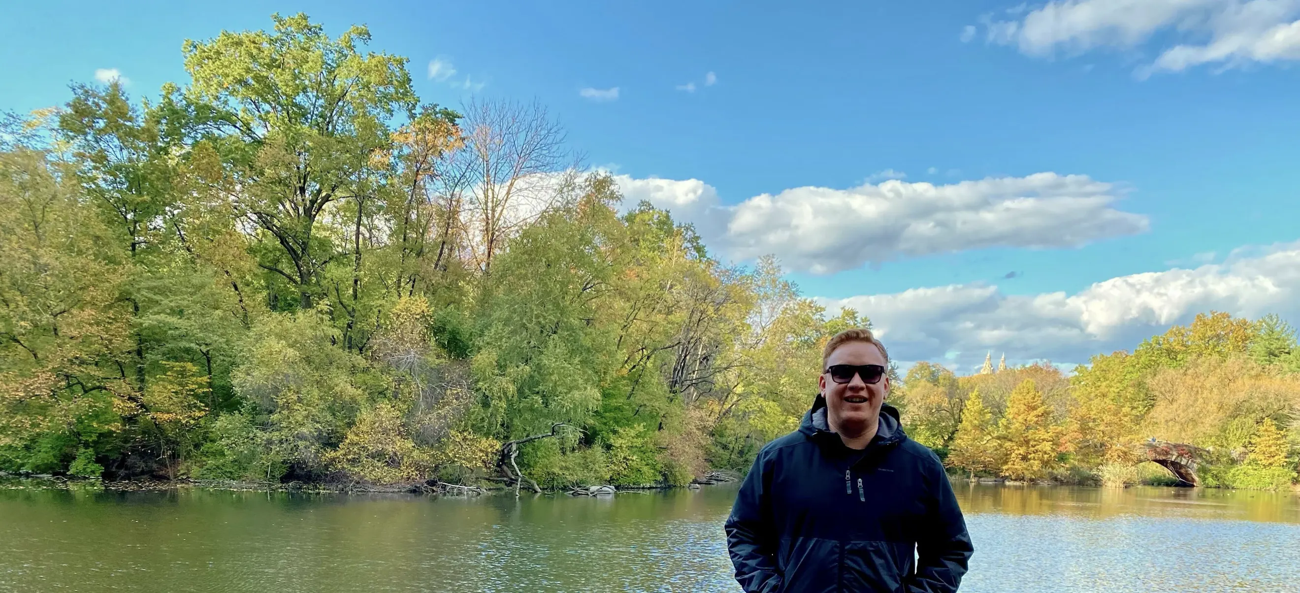 David stands by a lake, with trees and a blue sky with some clouds in the background.
