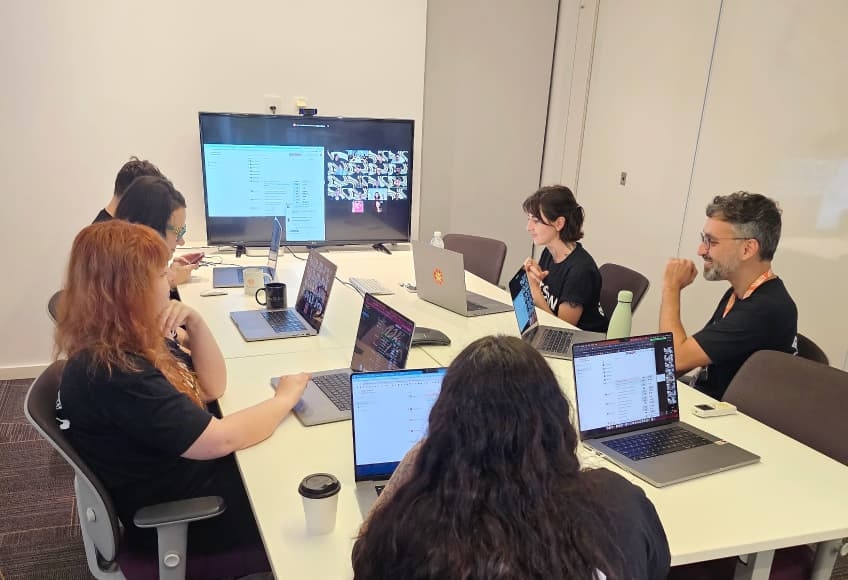 A team of six people seated at a table with laptops, attending an ArcTouch meeting with a large screen in a conference room.