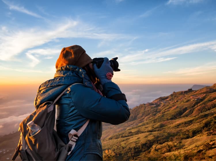 A person dressed in warm clothing takes a photograph of a mountainous landscape at sunrise or sunset. Clouds partially cover the lower hills, with a clear sky above.