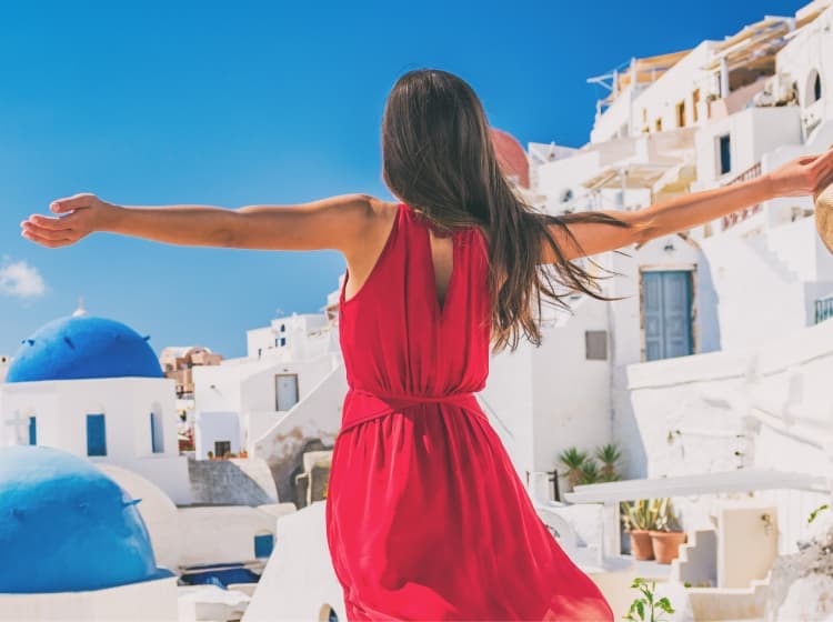 Person in a red dress with outstretched arms stands facing white buildings with blue domes under a clear blue sky, holding a hat in hand in a coastal town.