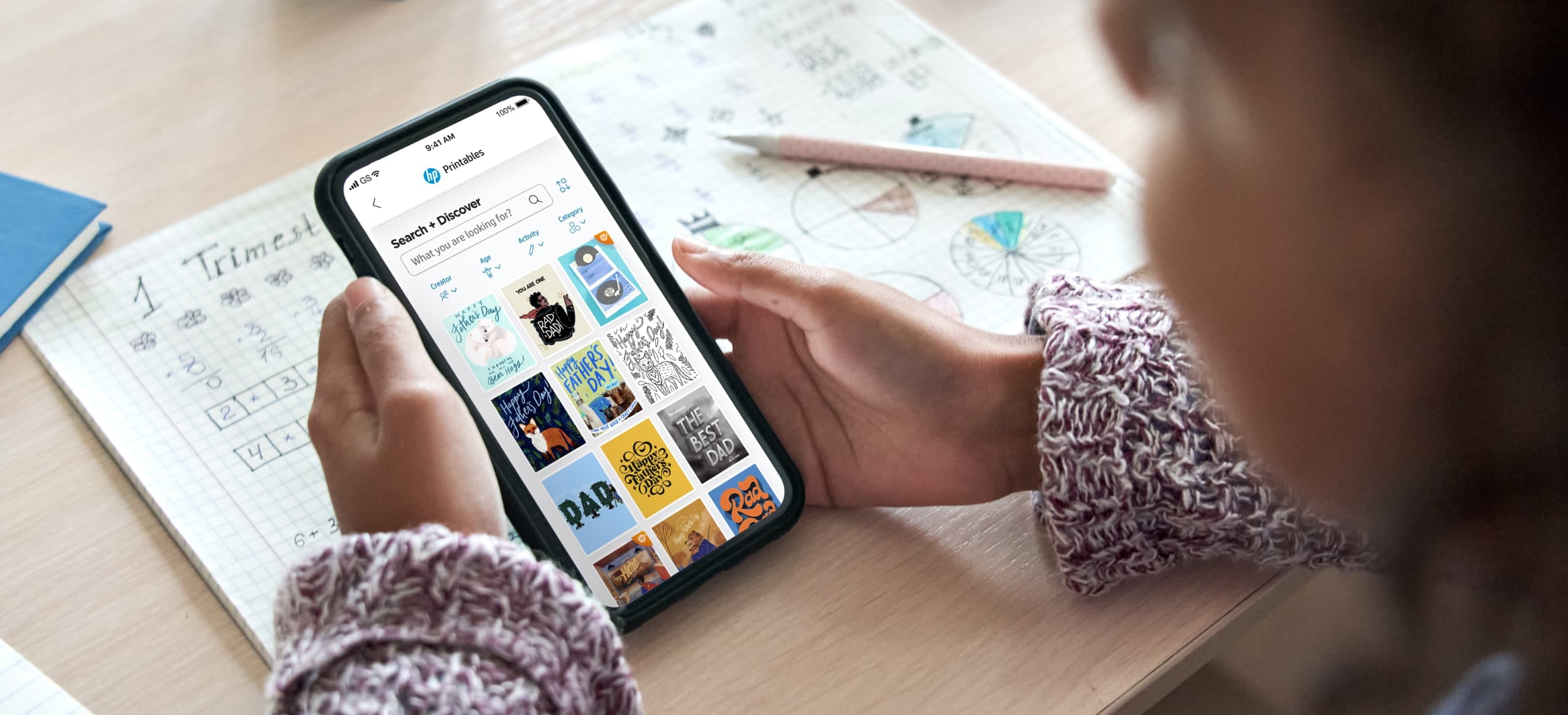 A person holds a smartphone displaying an HP Printables app profile while seated at a desk with paper, a pen, and additional documents or notebooks.