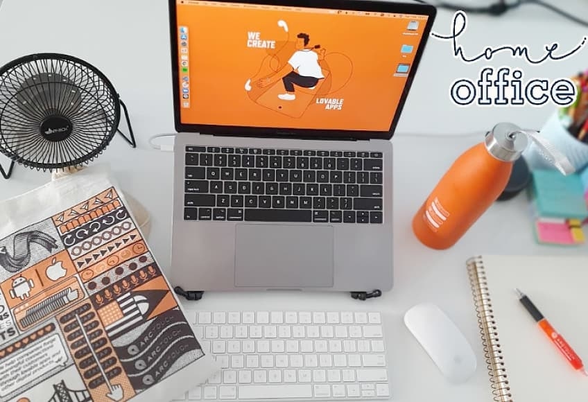 A laptop displaying an orange ArcTouch screen is on a desk with a small fan, notebook, orange water bottle, mouse, and tote bag. The text "home office" is visible in the background.