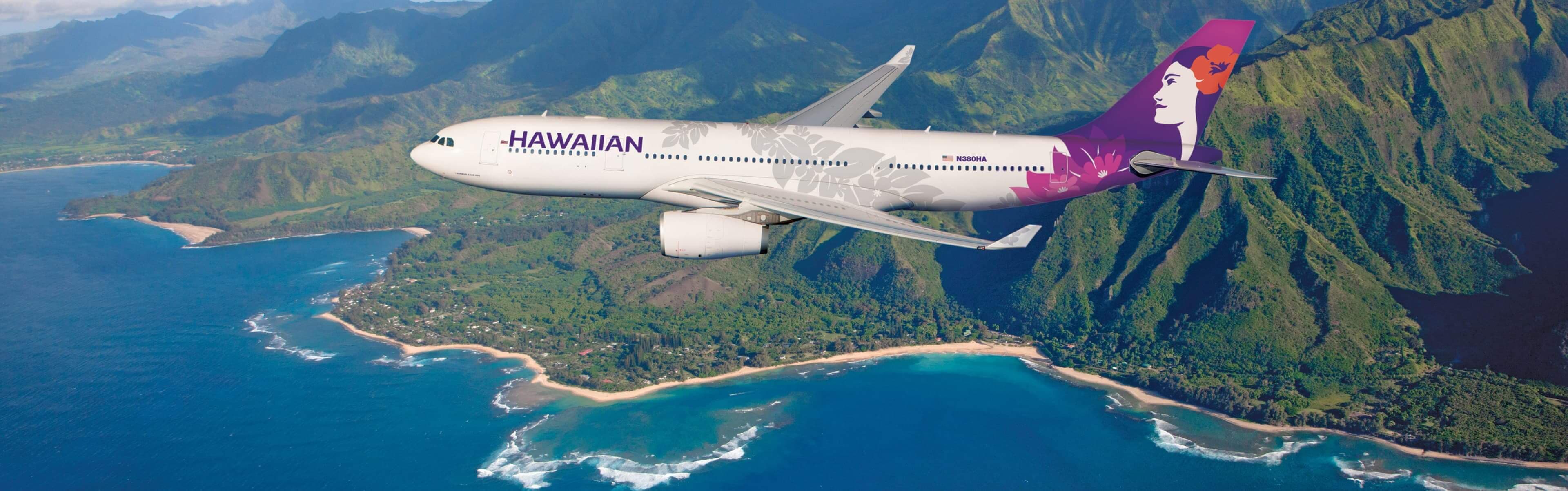 A Hawaiian Airlines airplane flies over a scenic coastline with lush green mountains and blue ocean waters