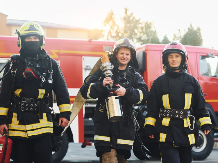 Three firefighters in full gear stand in front of a red fire truck.