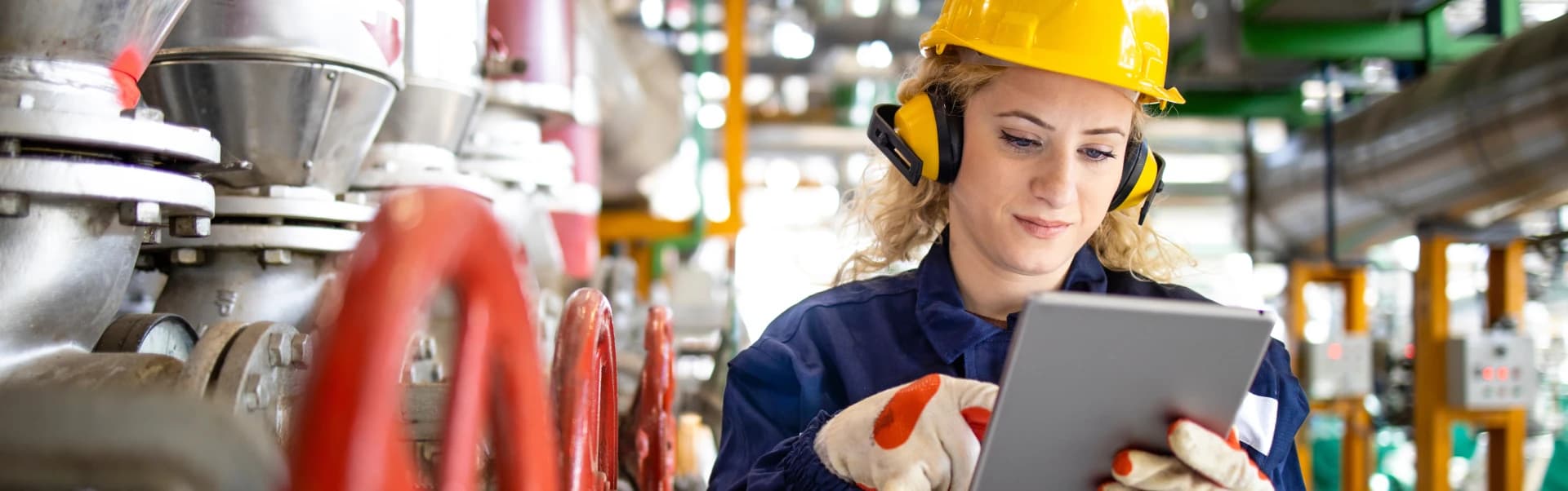 A woman dressed in protective work clothing operates a tablet in an industrial environment.